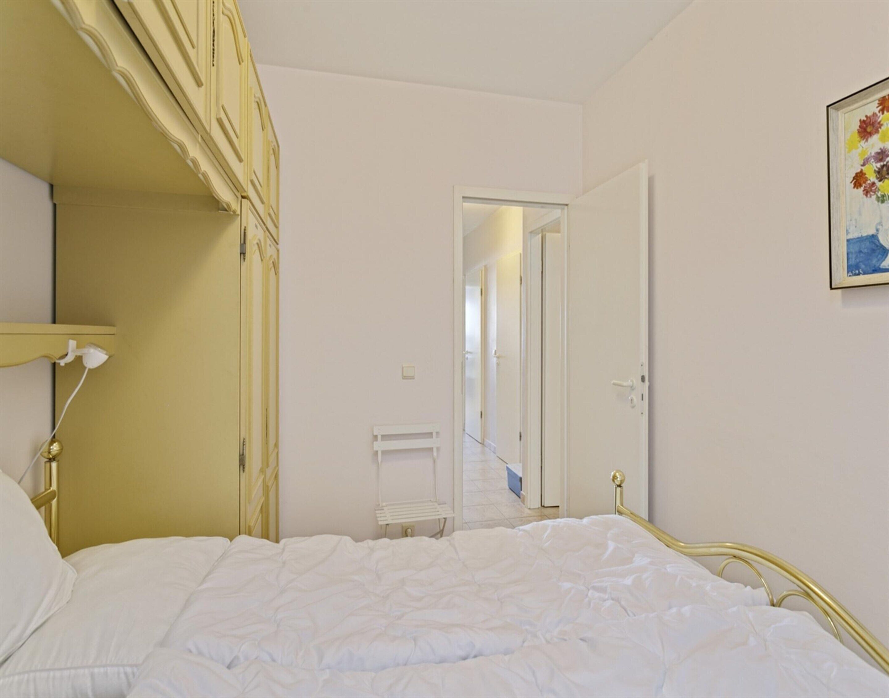 A bedroom with a bed, a bedside table, a lamp, and a door leading to another room.