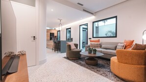 Una sala de estar moderna con un sofá, sillones y una mesa baja. Se observa un comedor en el fondo con una barra y sillas.