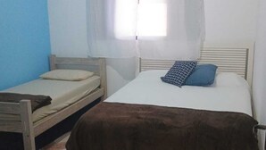 Eine kleine, einfache Unterkunft mit einem Bett, einem Nachttisch, einem fenster mit Vorhängen und einem gemusterten Boden.