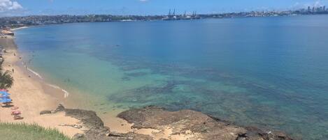 A coastal beach with clear water, a sandy shore, and a distant city skyline.