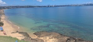 A coastal beach with clear water, a sandy shore, and a distant city skyline.