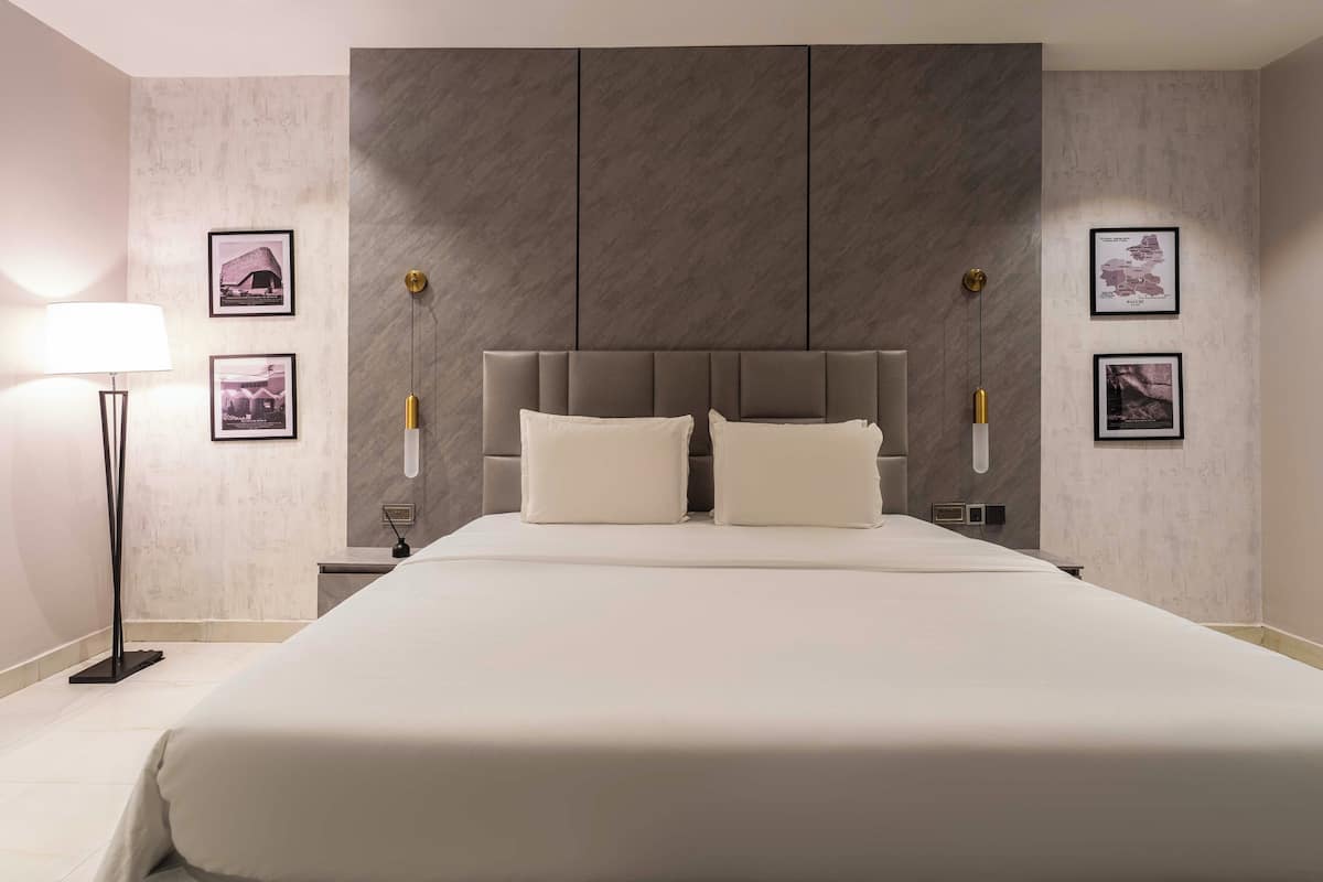 A hotel room with a large bed, a modern headboard, two bedside tables, a floor lamp, and framed pictures on the walls.