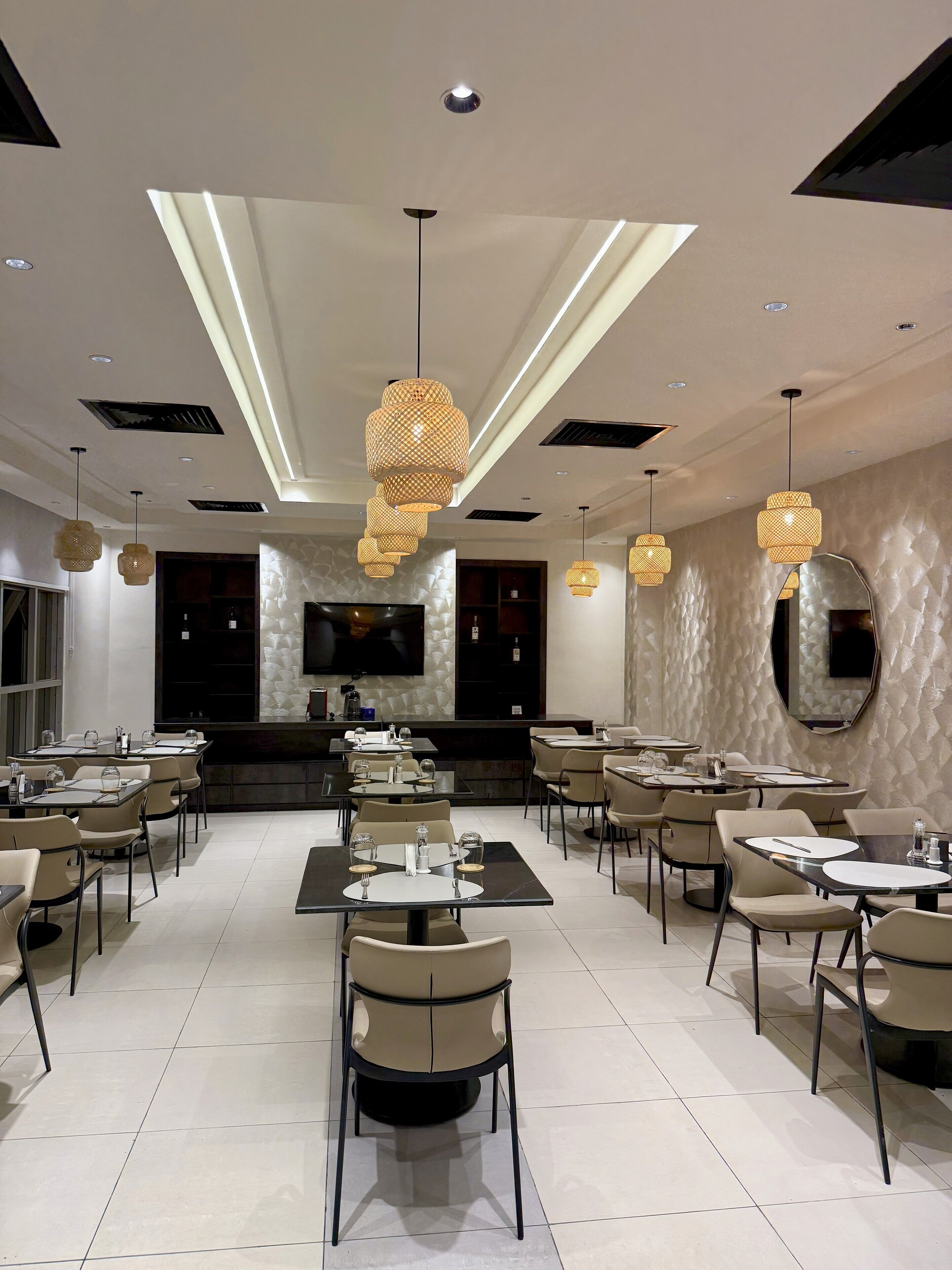 A modern dining area with multiple tables and chairs, a large flat-screen TV, and decorative woven light fixtures.
