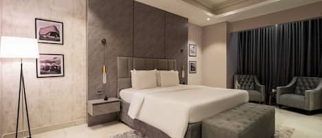 Un dormitorio moderno con una cama grande, una mesita de noche, un reposapiés, dos sillones y una cortina oscura.