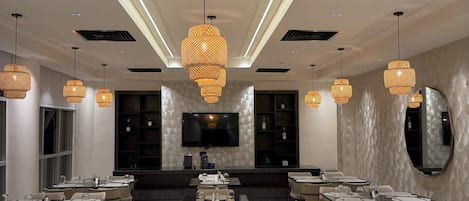Un'area pranzo moderna con diversi tavoli e sedie, un grande schermo televisivo e lampade decorative.