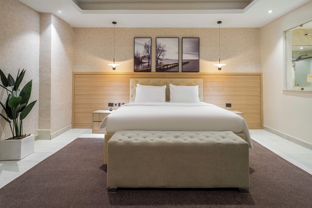 A modern bedroom with a large bed, a bedside table, a nightstand, a potted plant, and a wall-mounted artwork.