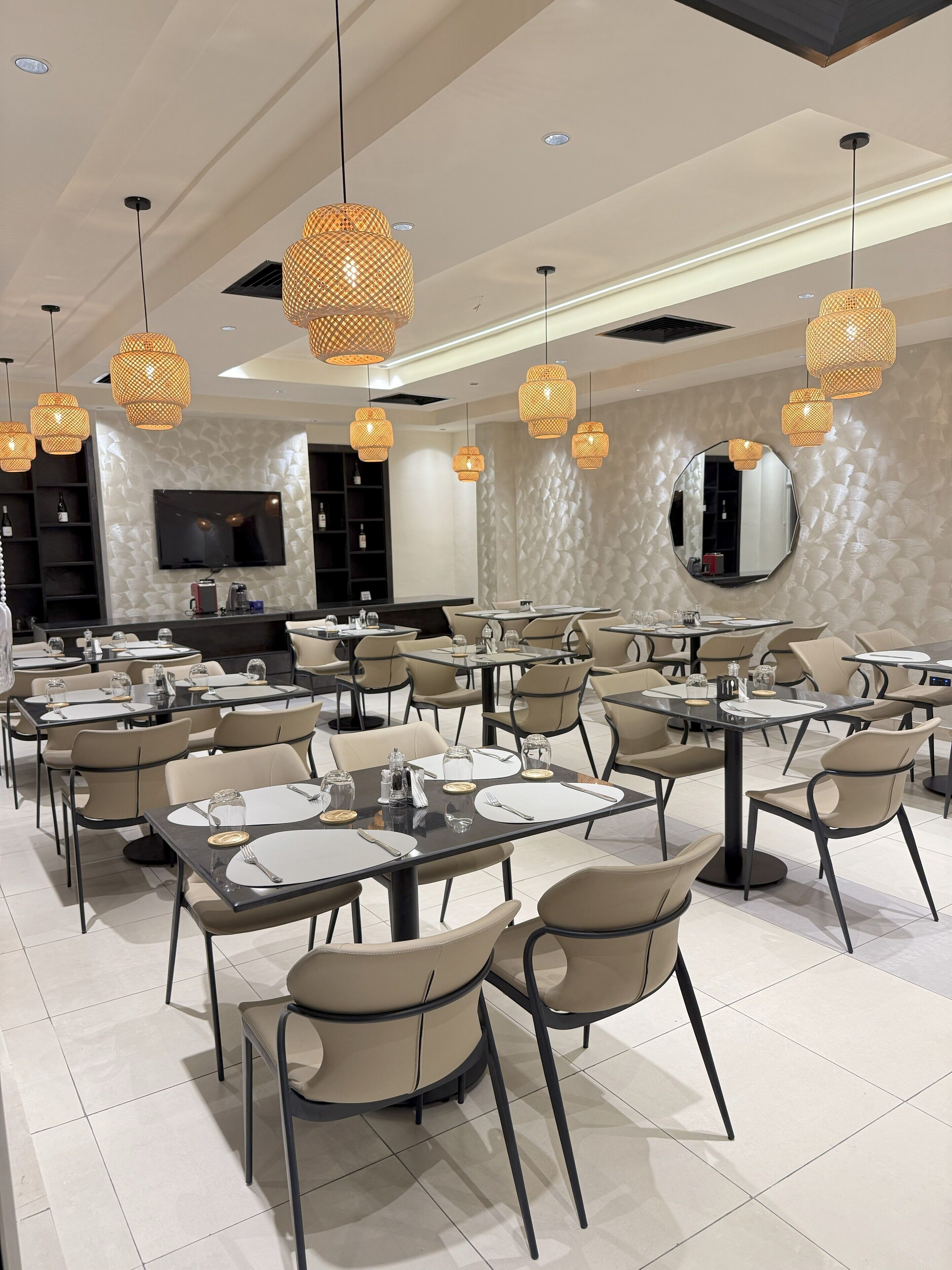A modern dining area with multiple tables and chairs, overhead wicker pendant lights, a flat-screen TV, and a textured wall.