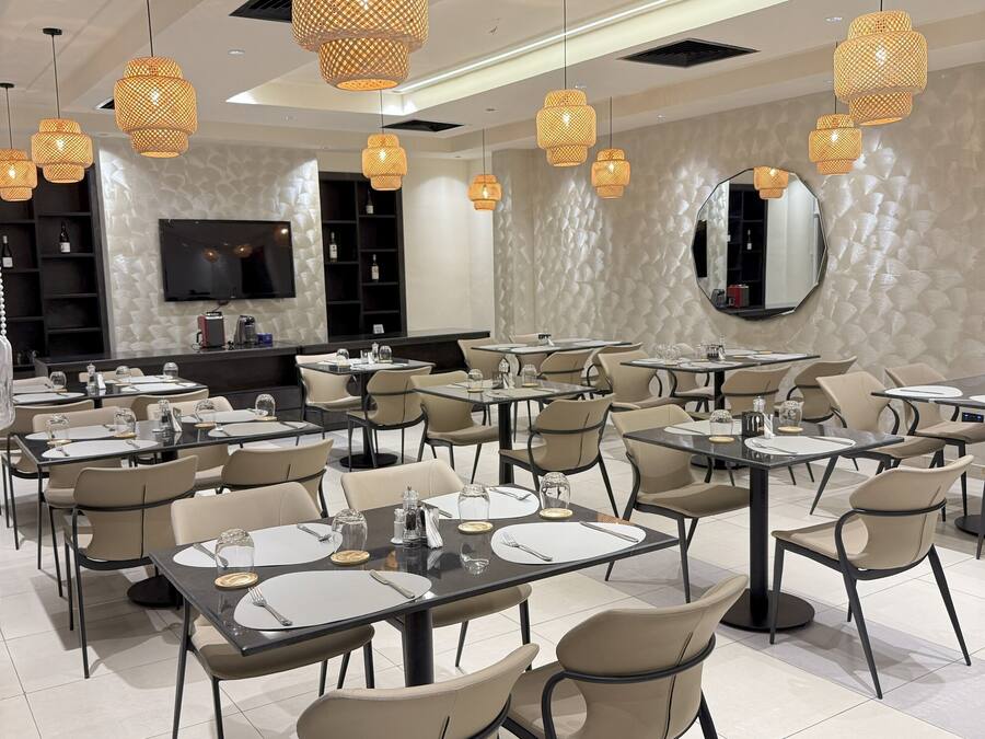A modern dining area with multiple tables and chairs, overhead wicker pendant lights, a flat-screen TV, and a textured wall.