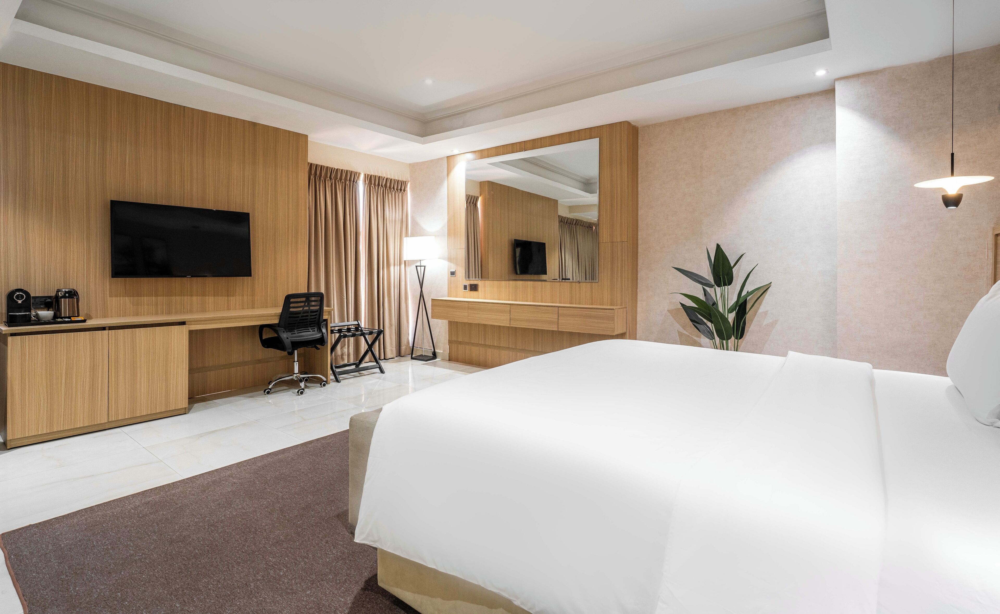 A hotel room with a large bed, a desk with a flat-screen TV, a chair, a coffee maker, and a potted plant.