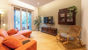 A living room with a sofa, a rocking chair, a television, a cabinet with plants, and a large window. - HouSmart Treves 2 (Bologna)