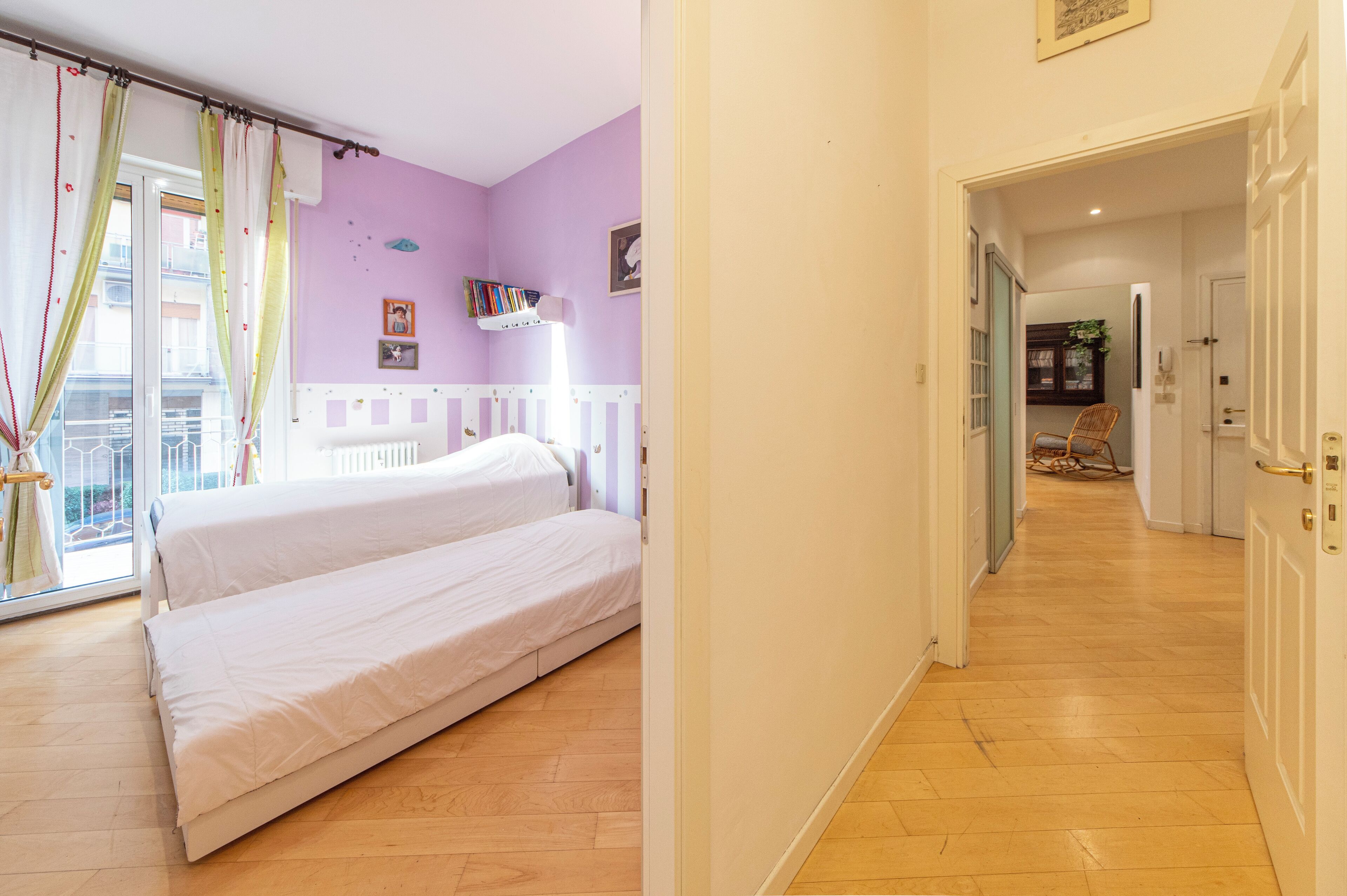 Un dormitorio con dos camas, una ventana con cortinas, una estantería y una puerta que da a otra habitación.
