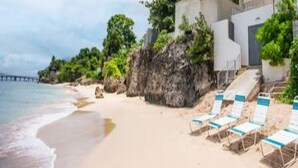 A beach with a rocky outcrop, a pier in the distance, and a building with a balcony overlooking the beach.