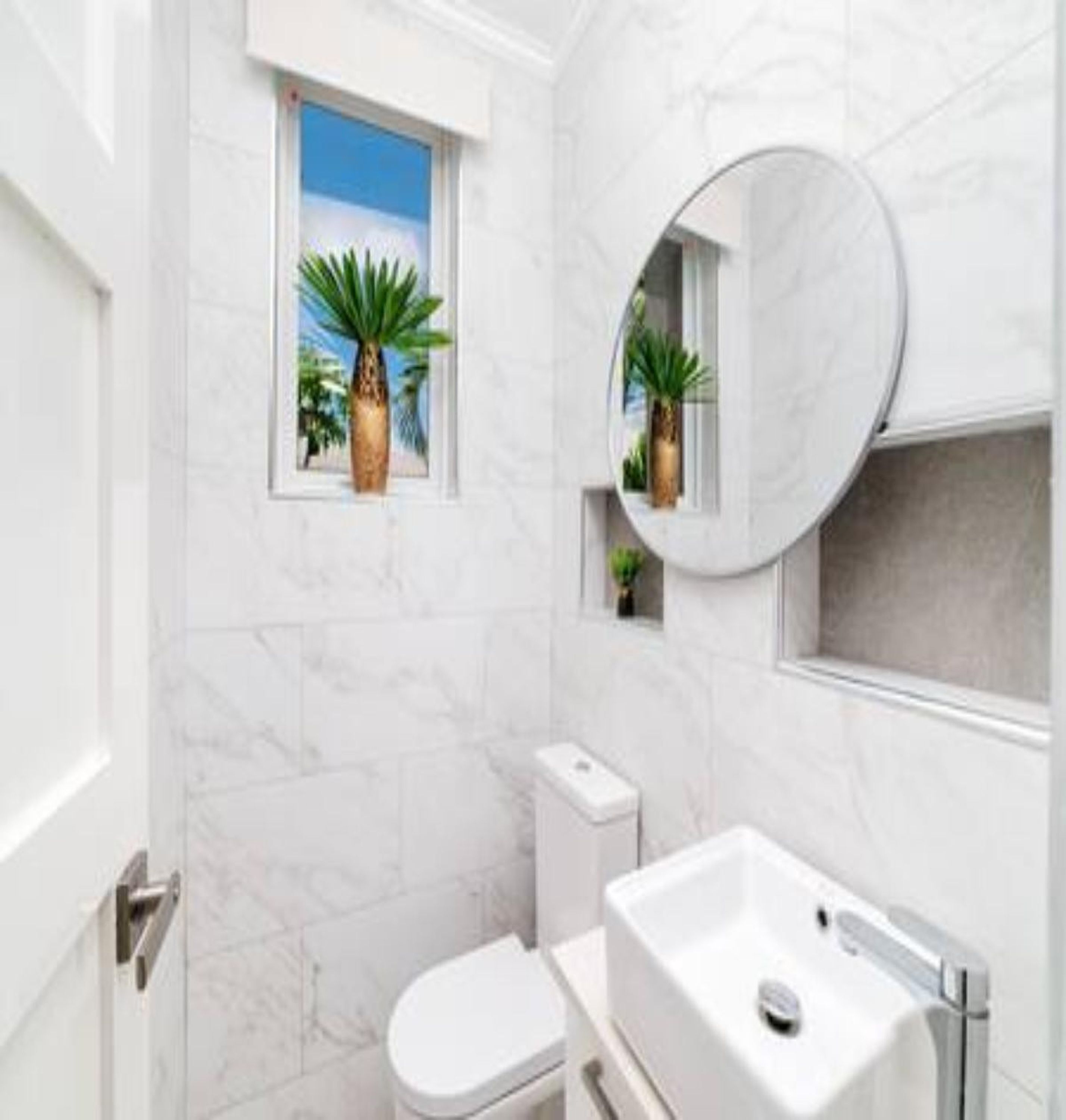 Une salle de bain équipée de toilettes, d’un lavabo et d’une fenêtre donnant sur des palmiers.