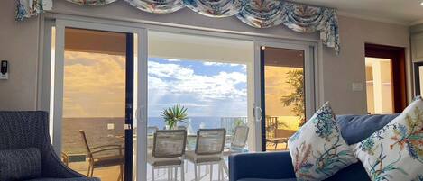 A room with a view of the sea, a balcony with chairs, and a sofa with a floral cushion.