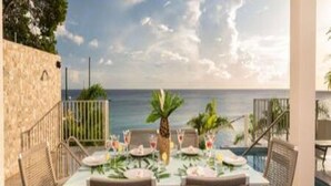 An outdoor dining area with a view of the ocean, featuring a table set for two with chairs, a stone wall, and a pool in the background.