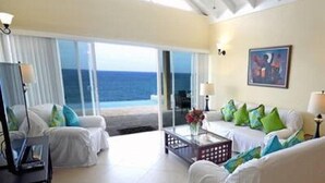 A spacious living room with a large window offering a view of the ocean, a sofa set, and a coffee table. - Je Suis Jamaica - 4 B R Beachfront Villa w Staff (Duncans)
