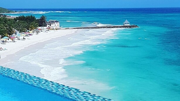 A beach resort with a swimming pool, beach umbrellas, and a lighthouse in the distance. - Je Suis Jamaica - 4 B R Beachfront Villa w Staff (Duncans)