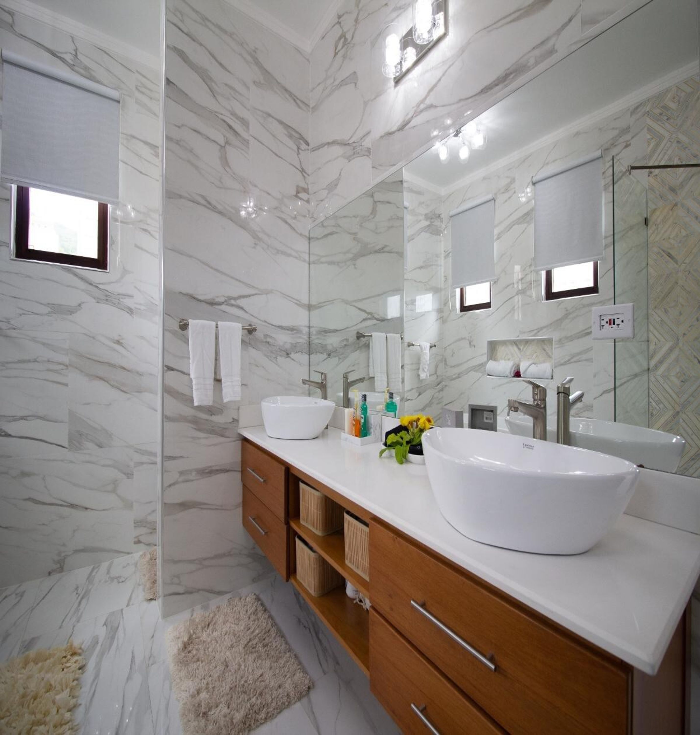 Un baño moderno con paredes de mármol, dos lavamanos, un espejo grande y una alfombra mullida.
