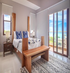 A modern bedroom with a large bed, a nightstand, a wooden bench, and a view of the ocean through large sliding doors.