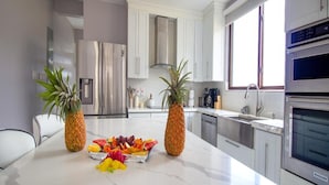 Cocina moderna con gabinetes blancos, electrodomésticos de acero inoxidable y una isla de mármol con fruta y dos plantas en macetas.