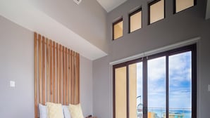 Un dormitorio moderno con cama, mesitas de noche, un banco, alfombra estampada y un gran ventanal corredero que da a un balcón con vistas al océano.