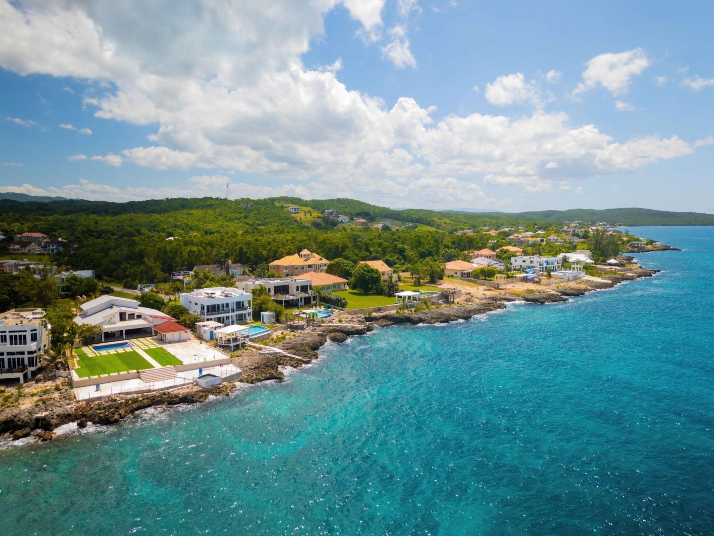 Eine Küstenwohngegend mit Häusern entlang der Küste, klarem blauen Wasser und grünen Hügeln im Hintergrund.