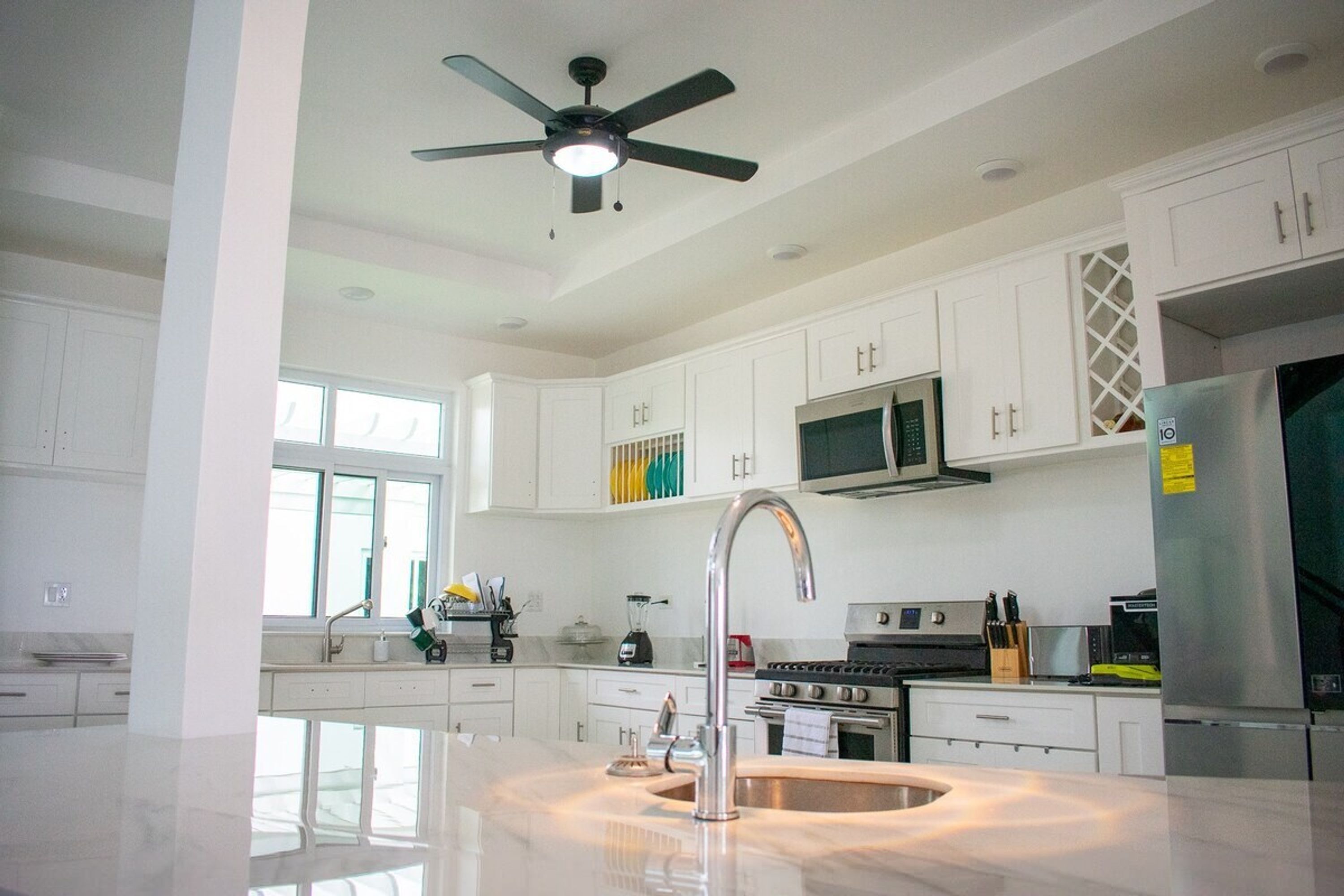 A modern kitchen with white cabinets, a stainless steel refrigerator, a built-in oven, and a ceiling fan.