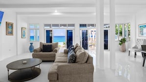 A spacious living room with a large window overlooking the sea, a beige sofa, a coffee table, and a potted plant. - The W Villa 5 B R Oceanfront w Staff (Whitehouse)