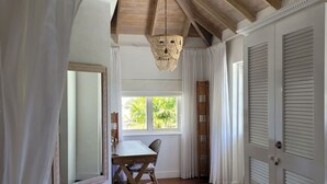 Una habitación con techo de madera, una lámpara de techo tejida, una mesa con sillas, un espejo grande y vistas al exterior con vegetación.