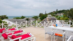 Eine Terrasse mit weißer Möbel, darunter Stühle und ein Tisch, mit Blick auf die Küste, im Hintergrund Häuser.