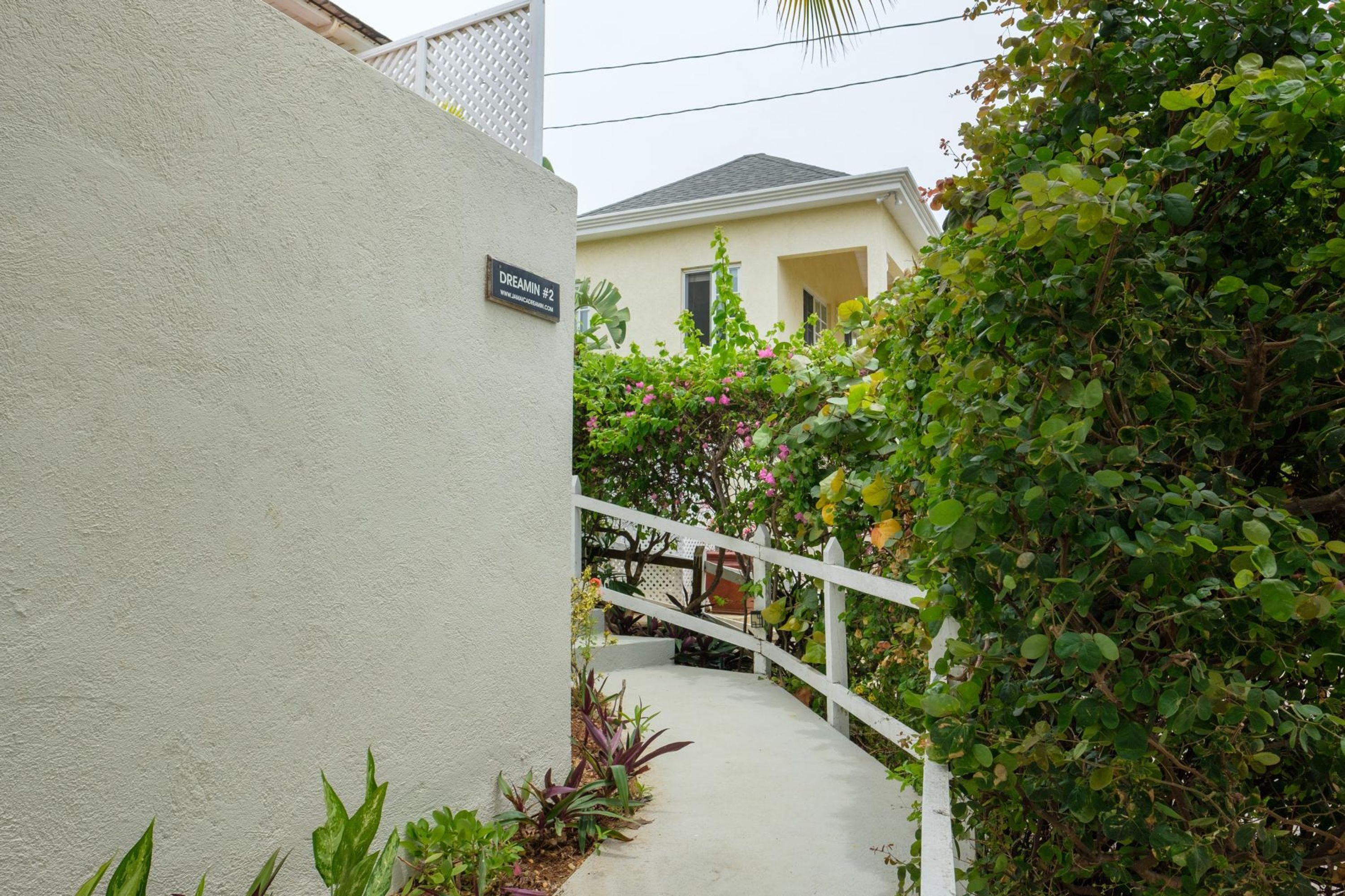 A white exterior wall with a sign reading "DREAMIN #2" and a white staircase leading to a house surrounded by lush greenery.