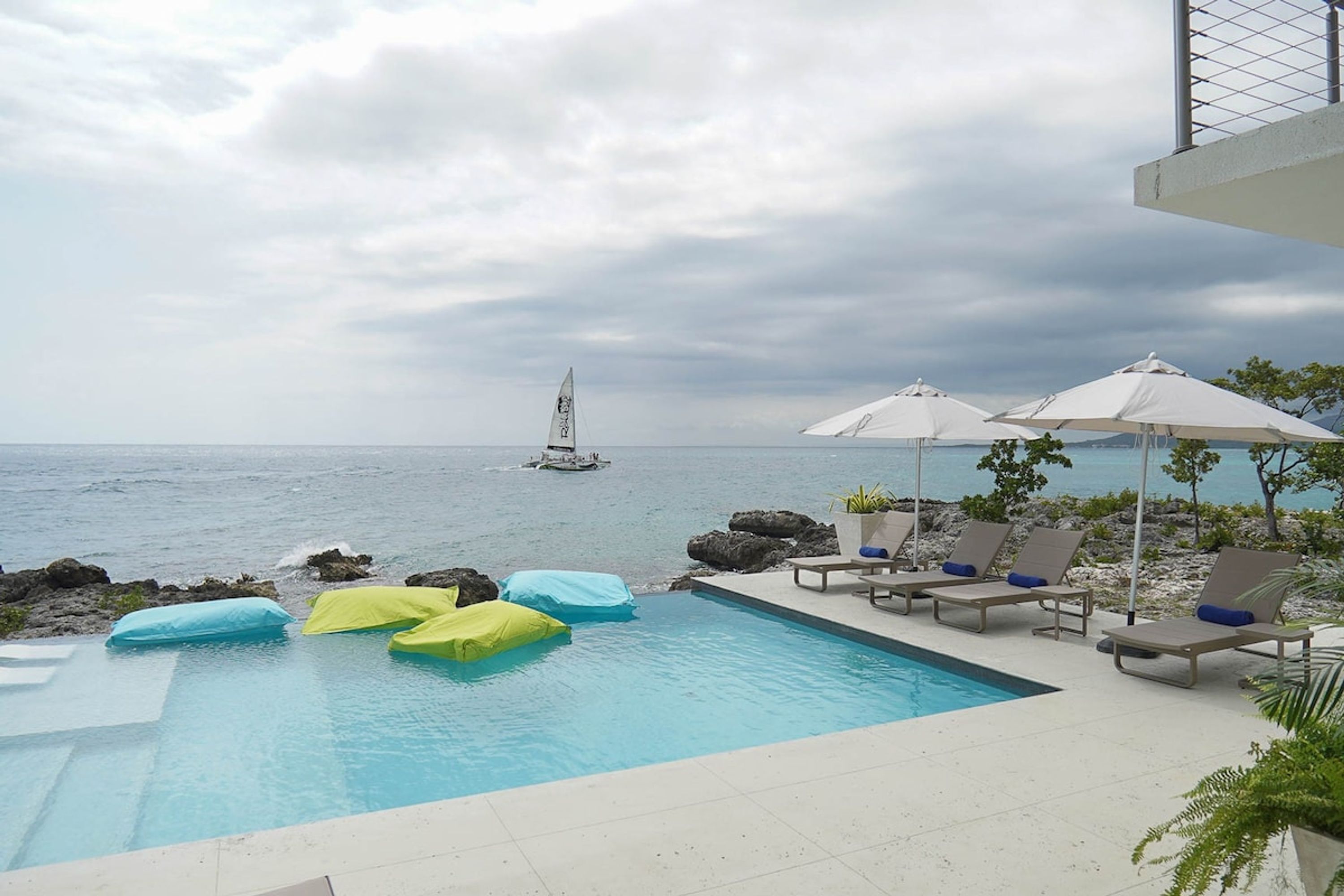 A pool with lounge chairs and umbrellas, a sailboat in the distance, and a view of the sea.