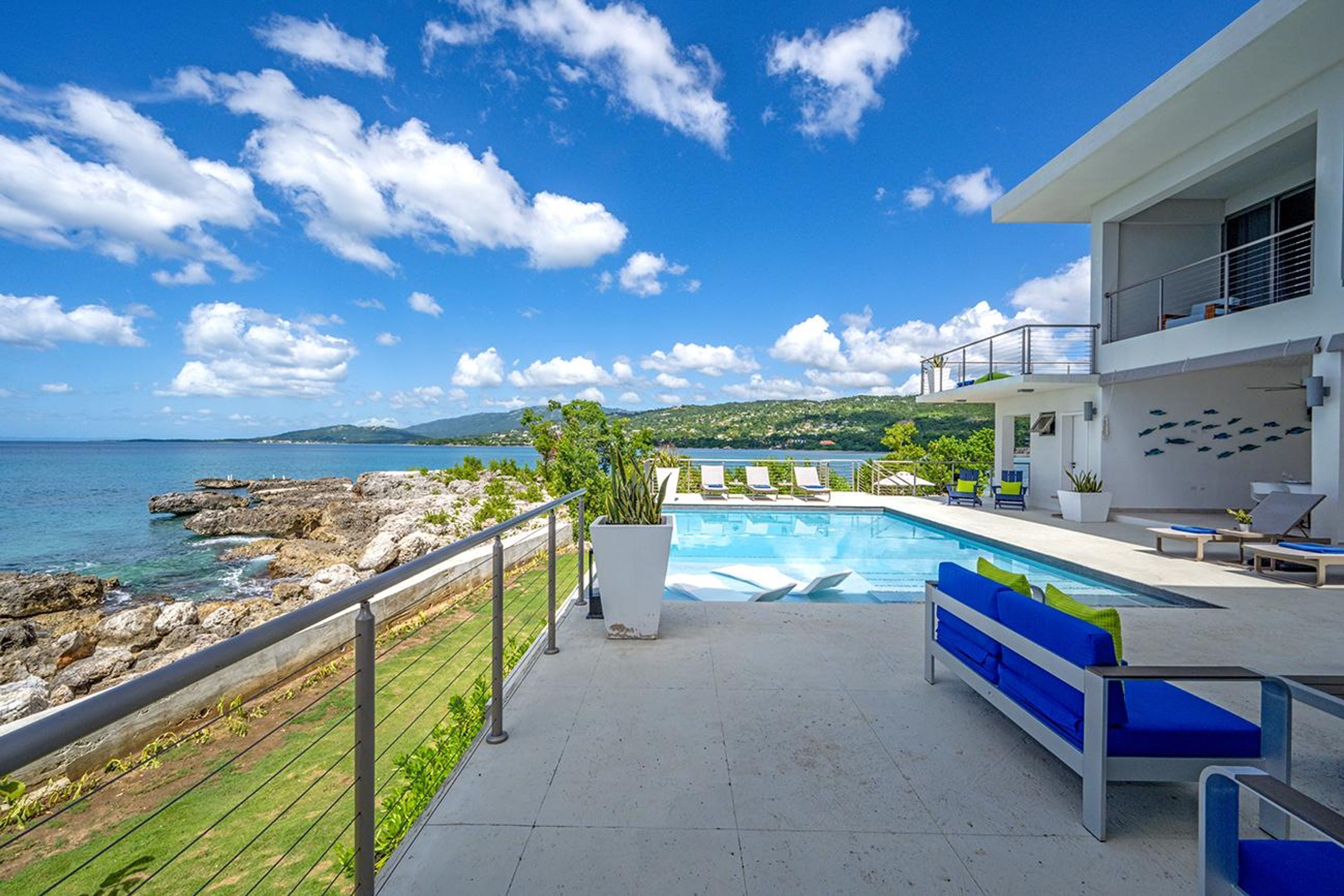 Un área de piscina moderna con vista al mar, un balcón con sillones de descanso y un edificio blanco con balcón.
