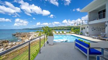 Un área de piscina moderna con vista al mar, un balcón con sillones de descanso y un edificio blanco con balcón.