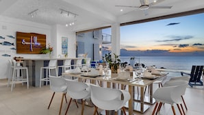 Un espace de restauration moderne avec vue sur la mer, des chaises blanches et un coin bar agrémenté d’une enseigne en bois.