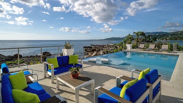 Un espace aménagé au bord de la piscine, avec des chaises longues et une table, offrant une vue sur une mer d’un bleu limpide et une côte rocheuse.