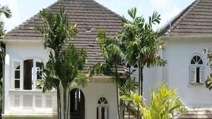 Una casa de dos pisos con techo de tejas, un porche con barandal blanco y un pequeño jardín con diversas plantas.