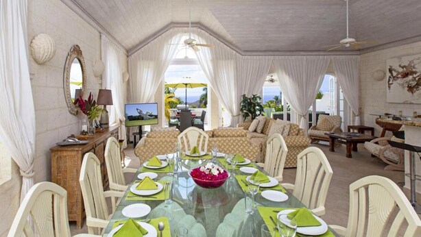 Un comedor con una mesa de vidrio preparada para seis personas, rodeada de sillas blancas. En el fondo, hay un sofá y un televisor.