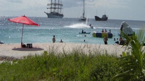 Una playa con una sombrilla roja, gente nadando y un barco grande al fondo.