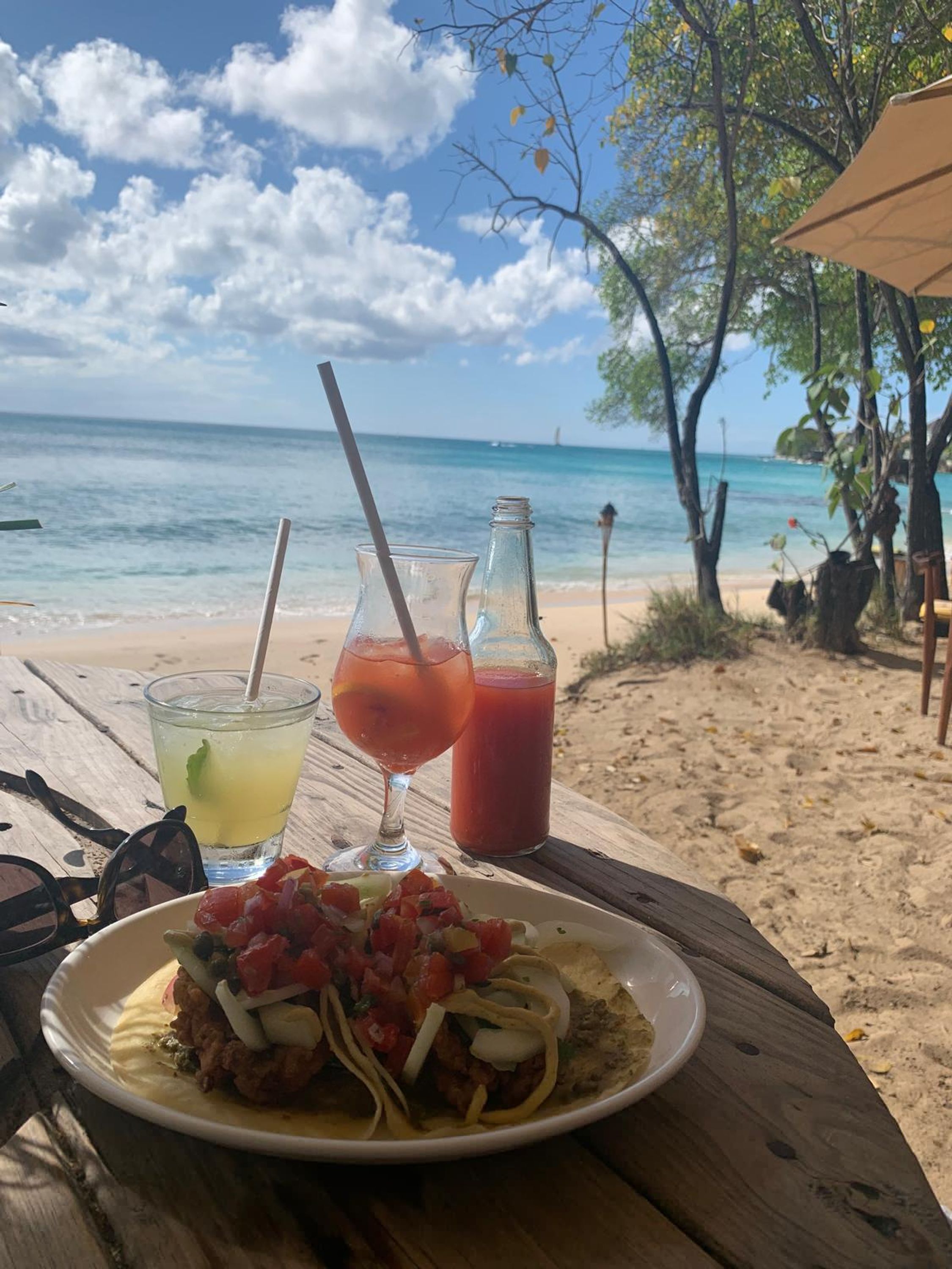 Una mesa en la playa con un plato de comida, dos bebidas y gafas de sol.