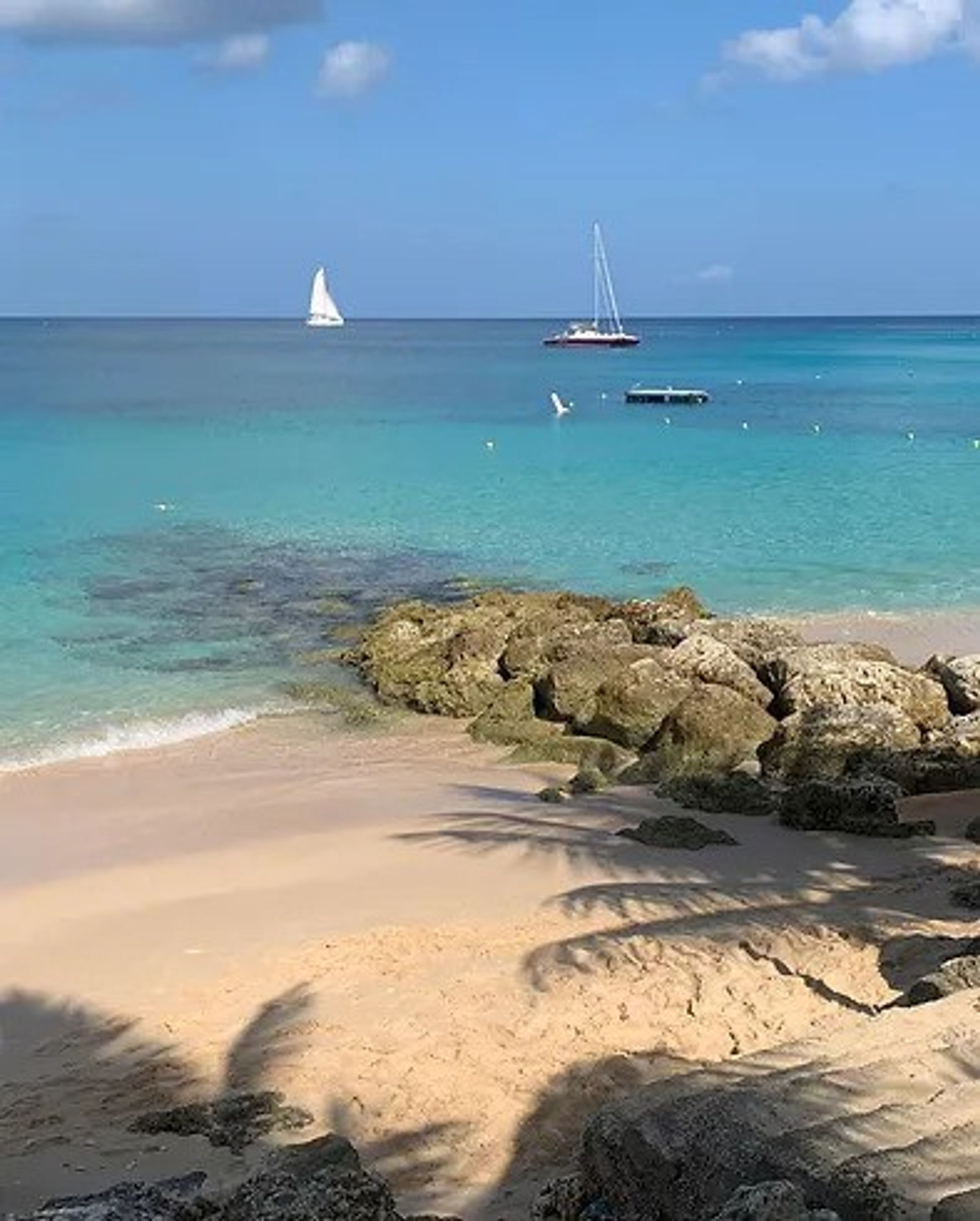 Una playa con agua turquesa cristalina, una costa rocosa y un velero a lo lejos.
