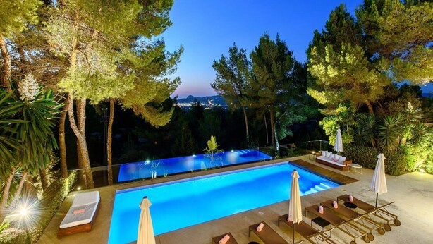 A swimming pool with lounge chairs and umbrellas, surrounded by trees and a view of the city at dusk.