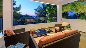 A modern outdoor seating area with wicker furniture, a glass balustrade, and a view of trees and hills.