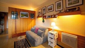 Ein gemütliches Zimmer mit einer Couch, einem kleinen runden Tisch und einer Heizung. An der Wand hängen Bilder in Rahmen und ein Regal mit dekorativen Gegenständen.