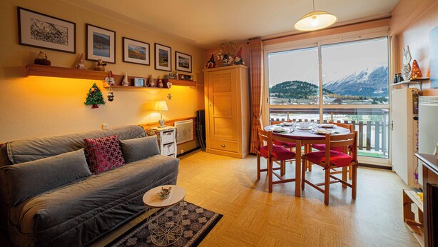 Ein gemütliches Wohnzimmer mit einer Couch, einem kleinen Tisch und einem Essbereich mit Stühlen. Ein Fenster bietet Blick auf Berge und es gibt einen Balkon.