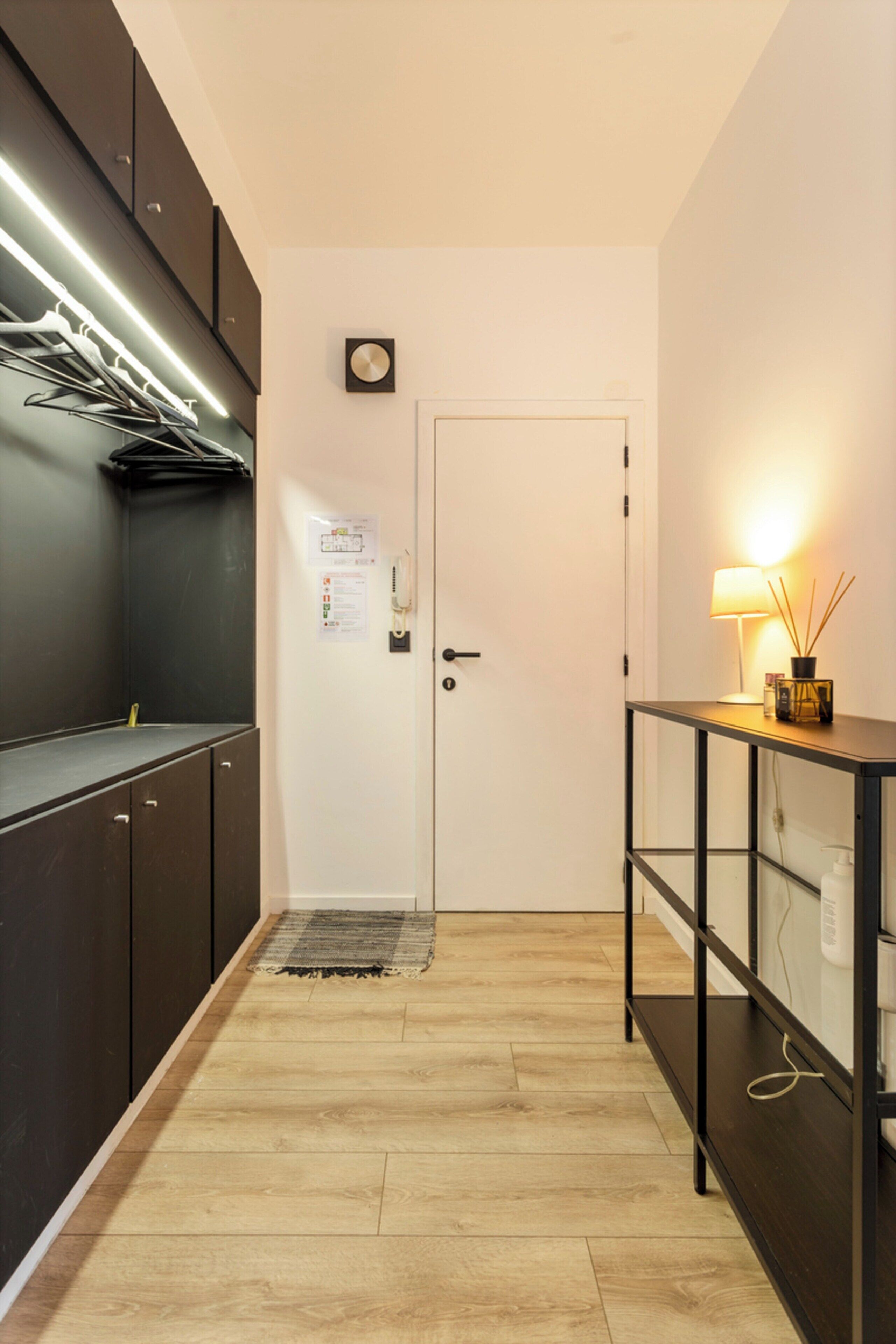 A hallway with a wooden floor, a door with a calendar, a lamp on a console table, and a coat rack.