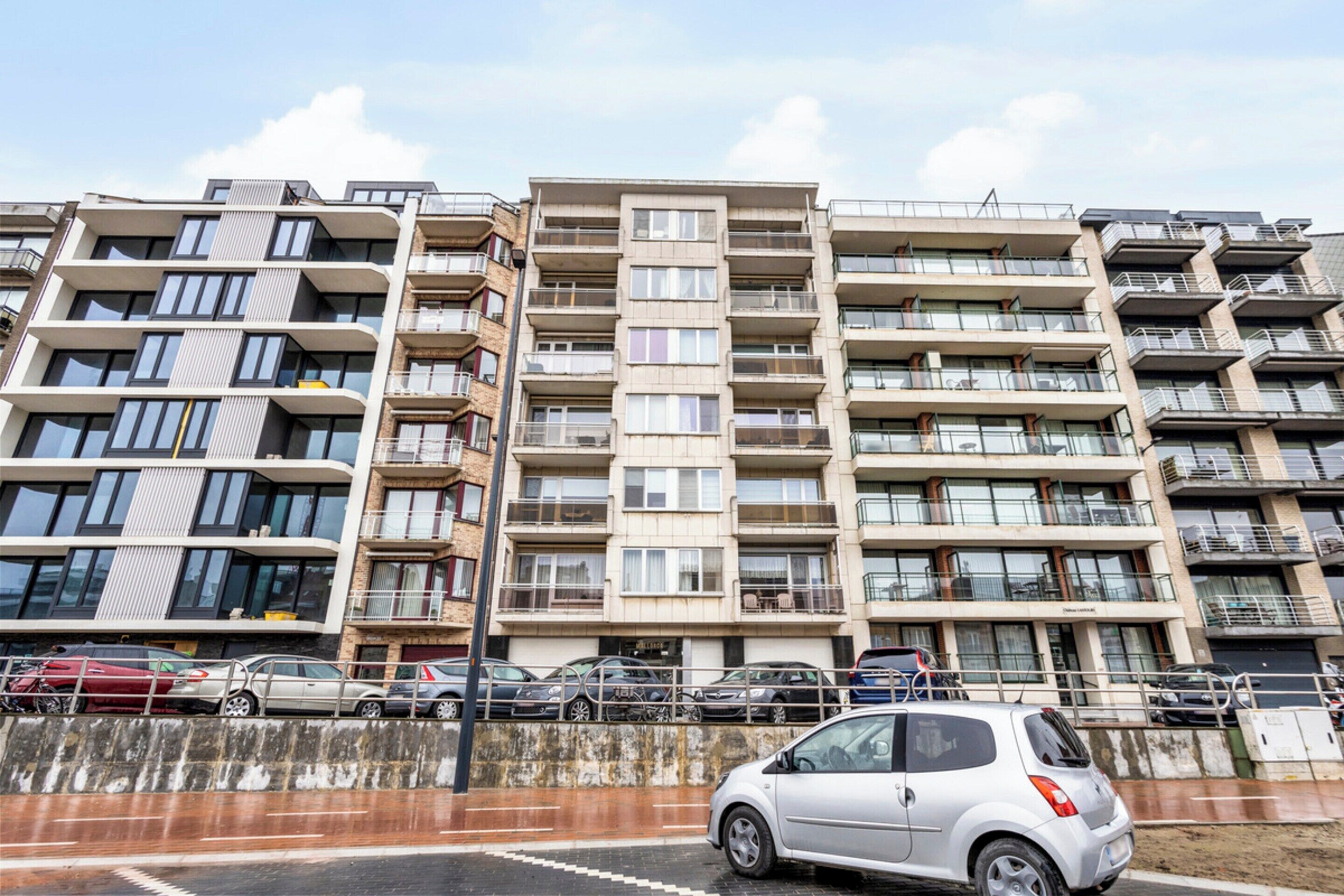 A multi-story residential building with balconies, a parking area, and a small car in the foreground.