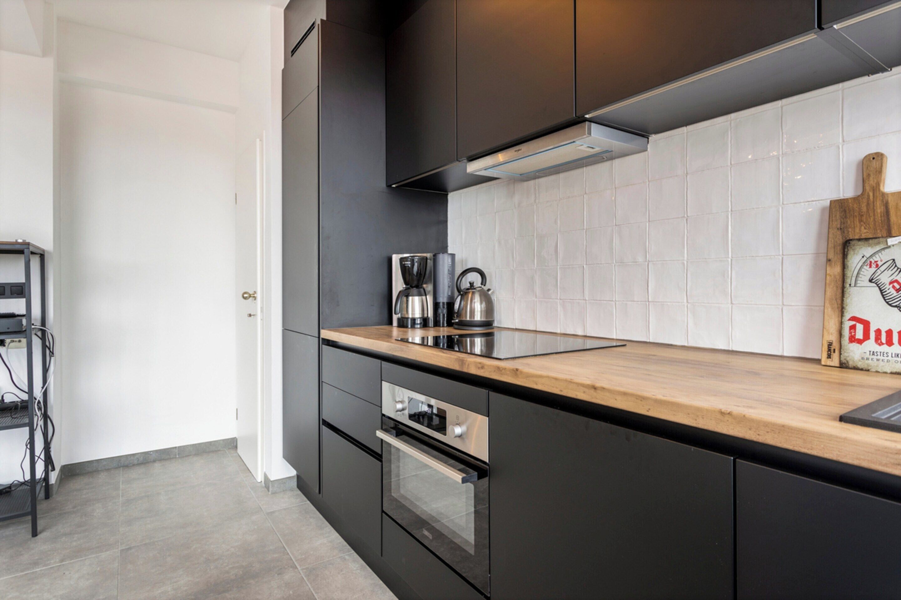 A modern kitchen with dark cabinetry, a wooden countertop, a kettle, a coffee maker, and a wooden cutting board with text.