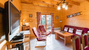 Interno accogliente di una baita in legno con TV, tavolo in legno, due poltrone a dondolo e un divano.