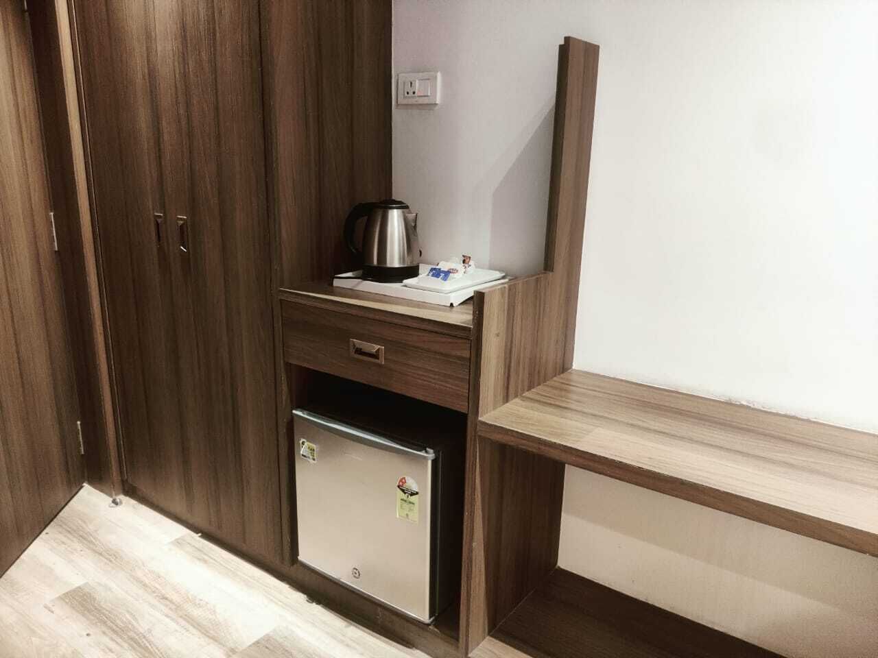 A compact room with a wooden wardrobe, a small refrigerator, a kettle, and a wooden bench.